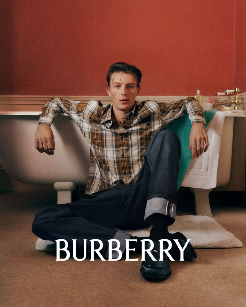 Man in plaid shirt and jeans seated on floor in front of bathtub in a casual fashion shot, showcasing Burberry logo at bottom of frame.