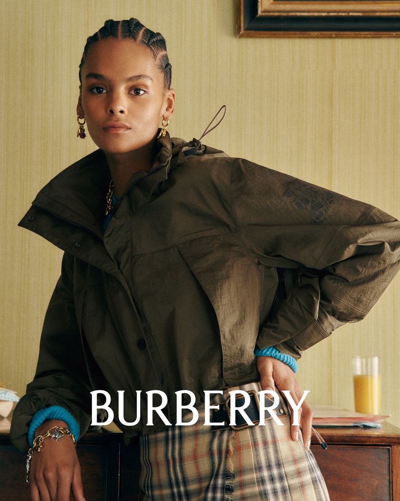 Fashionable young woman poses wearing a brown jacket and plaid skirt against a light-yellow wall in an indoor setting, displaying Burberry brand.