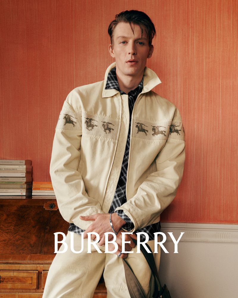 Young man in a light-beige Burberry jacket with horse graphics over a plaid shirt, posing indoors against a warm orange wall, fashion shoot.