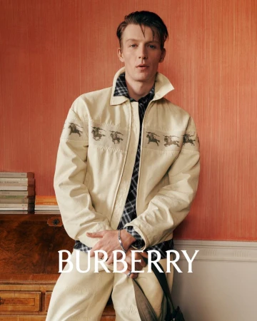 Young man in a light-beige Burberry jacket with horse graphics over a plaid shirt, posing indoors against a warm orange wall, fashion shoot.