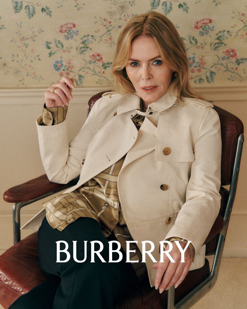 Mature woman in a beige trench coat posing in a chair against a floral wallpaper backdrop in a fashion editorial shot.