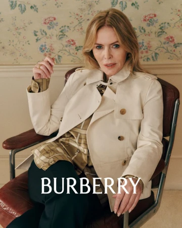 Mature woman in a beige trench coat posing in a chair against a floral wallpaper backdrop in a fashion editorial shot.