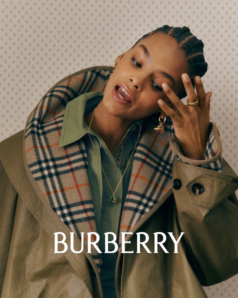 Fashion shot of an adult female model in a Burberry trench coat looking toward the camera in an indoor setting with patterned wallpaper.