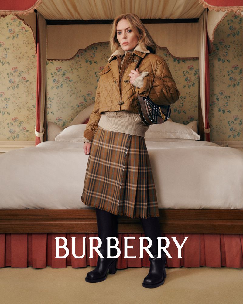 A woman in a brown jacket, sweater and kilt with a brown handbag stands in front of a bed with a floral headboard. Burberry logo in view.