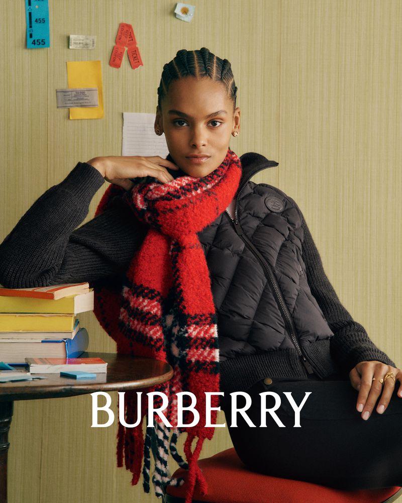 A woman with dark hair wearing a quilted jacket and a red plaid scarf poses indoors. She is seated at a table with books.