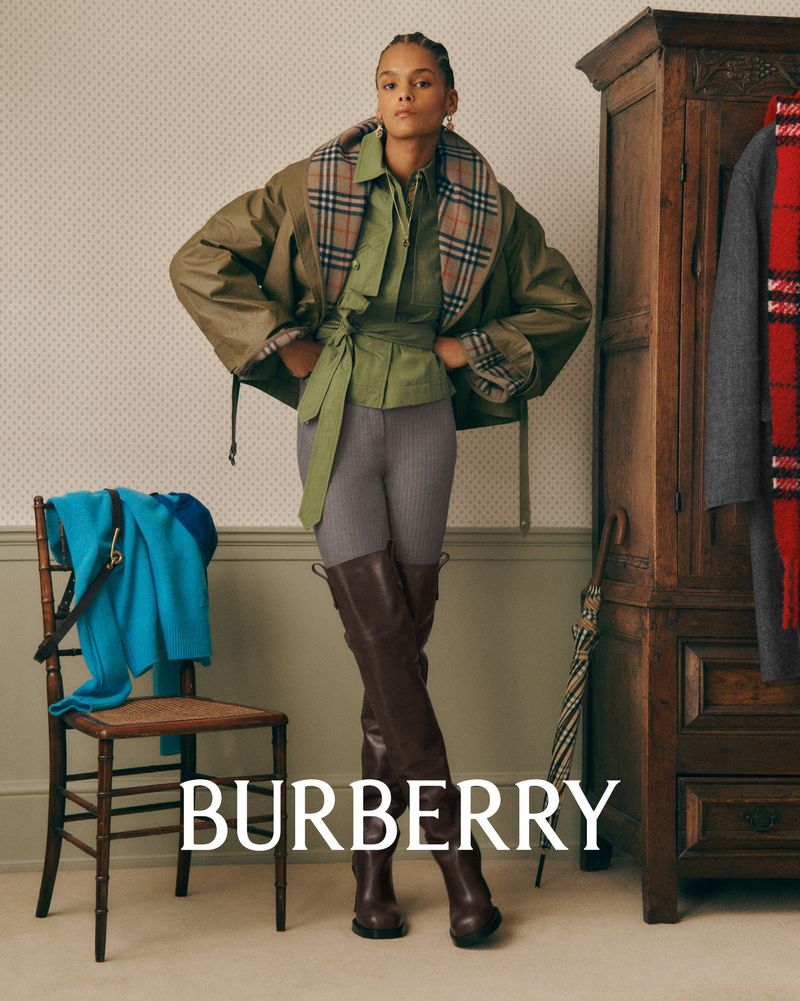 A woman stands indoors wearing a green outfit and brown boots, with a Burberry coat draped over her shoulders and accessories around her.