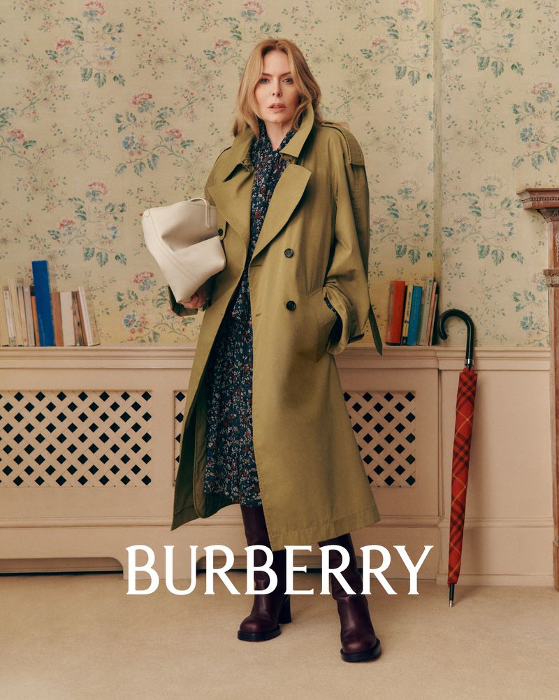 An adult woman with a handbag and wearing a trench coat stands in an indoor setting with wallpaper and books in a fashion shot.