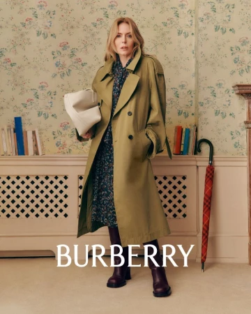 An adult woman with a handbag and wearing a trench coat stands in an indoor setting with wallpaper and books in a fashion shot.