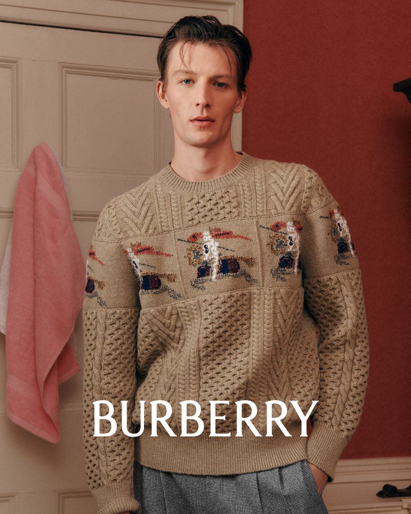 Adult male model wearing a Burberry patterned knit sweater in an indoor setting with soft lighting and a neutral color palette.