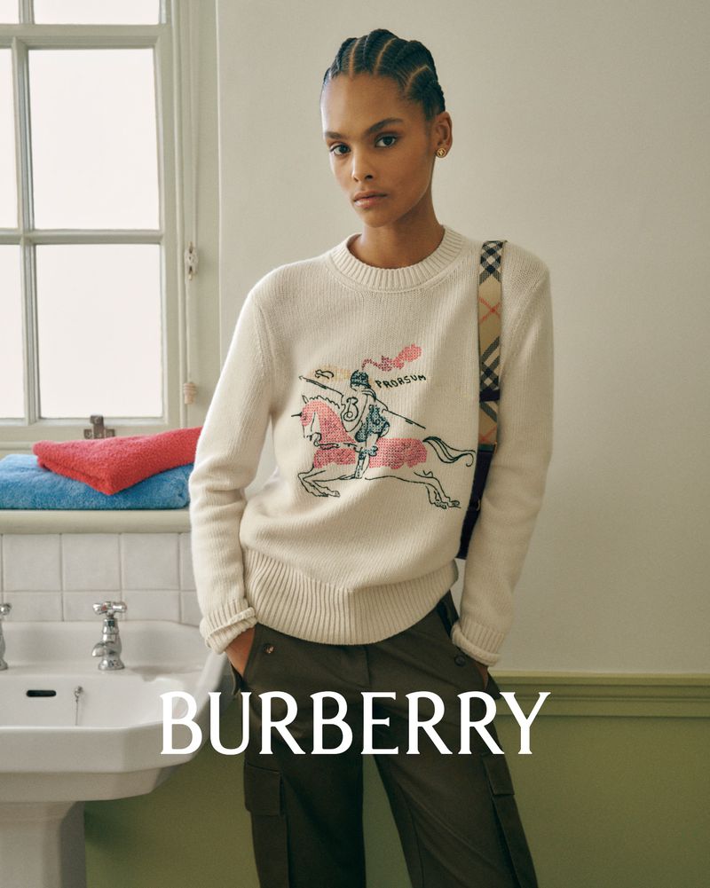 Fashion shot of a Black female model wearing a cream sweater with a knight graphic, posing in a bathroom with a Burberry bag.