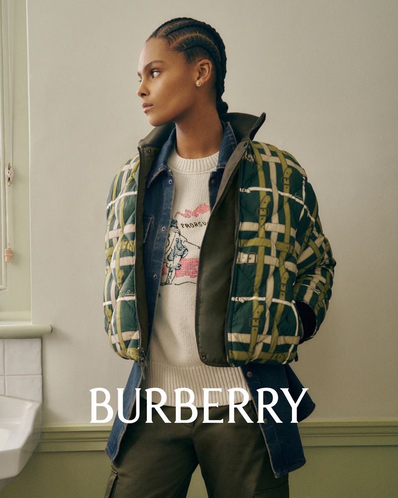 A woman in a Burberry jacket, denim shirt, and light sweater poses indoors in front of a wall with a white sink.