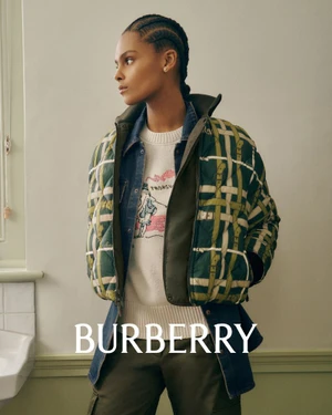 Model with braided hair wearing a green plaid jacket over a white sweater against an off-white wall
