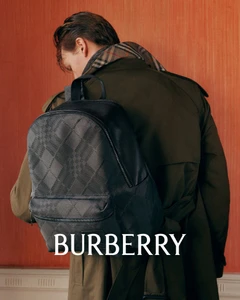 Man wearing a green trench coat and checked scarf carrying a dark patterned backpack against an orange wall.