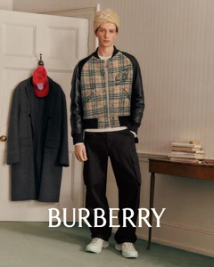 Model wearing a plaid and leather bomber jacket with a beanie standing in a room with a grey coat on a door and stacked books on a table