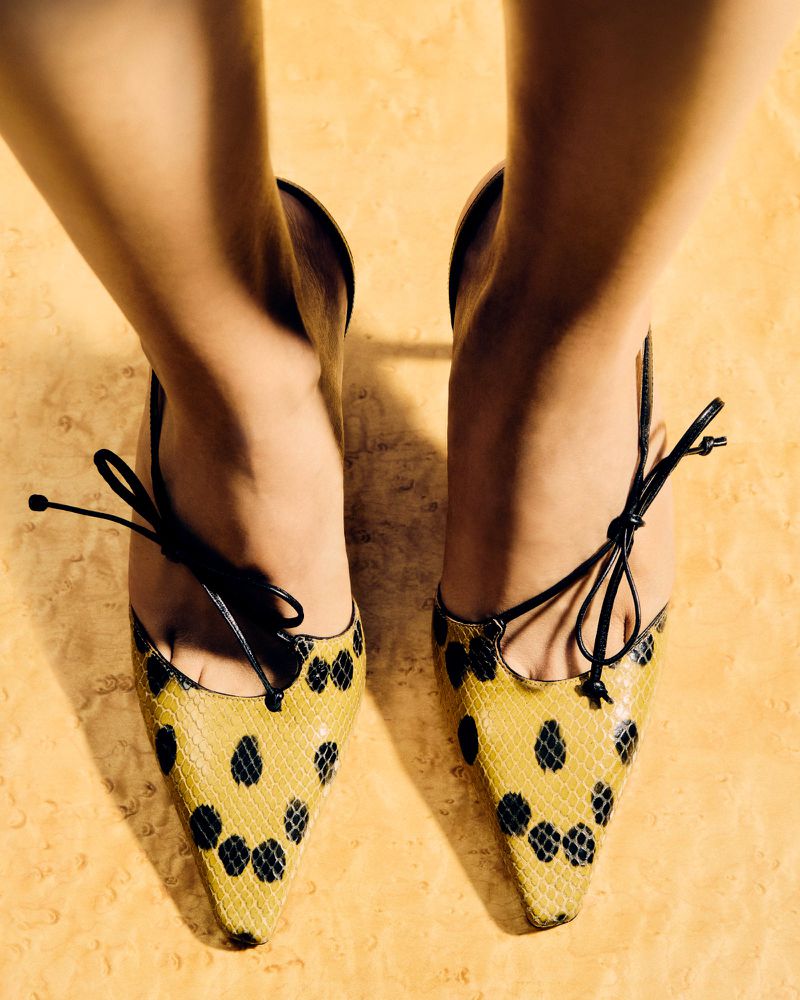 Close-up top shot of a pair of women's high heels with a black spotted yellow snakeskin pattern. Ankle straps are tied with black ribbons.
