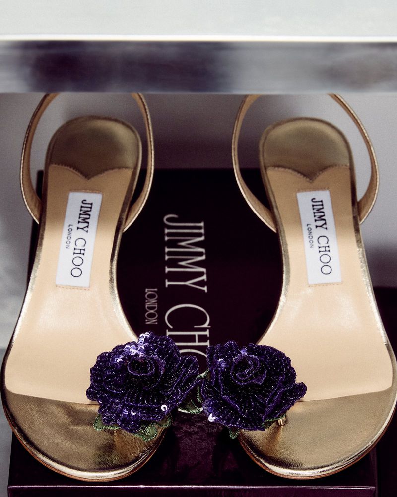 Gold Jimmy Choo sandals with purple floral embellishments displayed on a shelf, presented in an eye-level, medium studio shot.