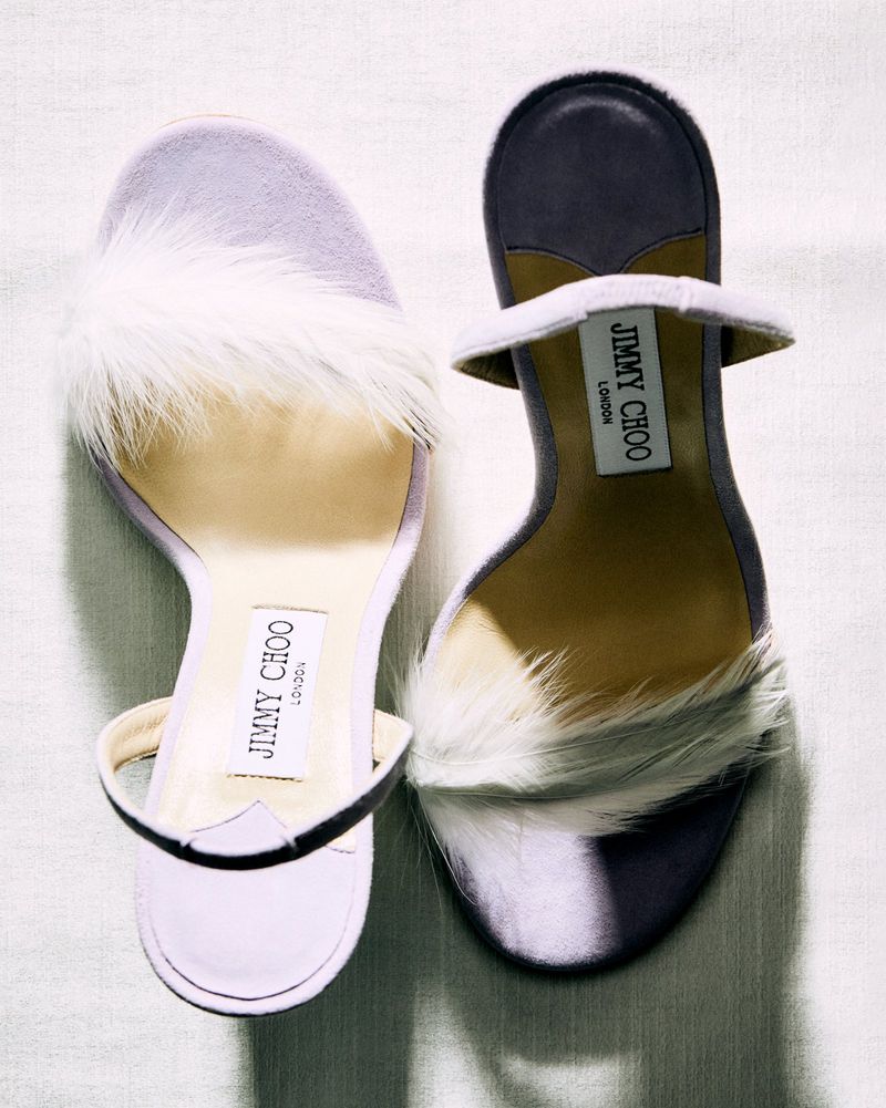 Overhead shot showcases two Jimmy Choo heeled sandals with white feather detailing, presented on a textured off-white surface.