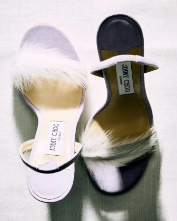 Overhead shot showcases two Jimmy Choo heeled sandals with white feather detailing, presented on a textured off-white surface.