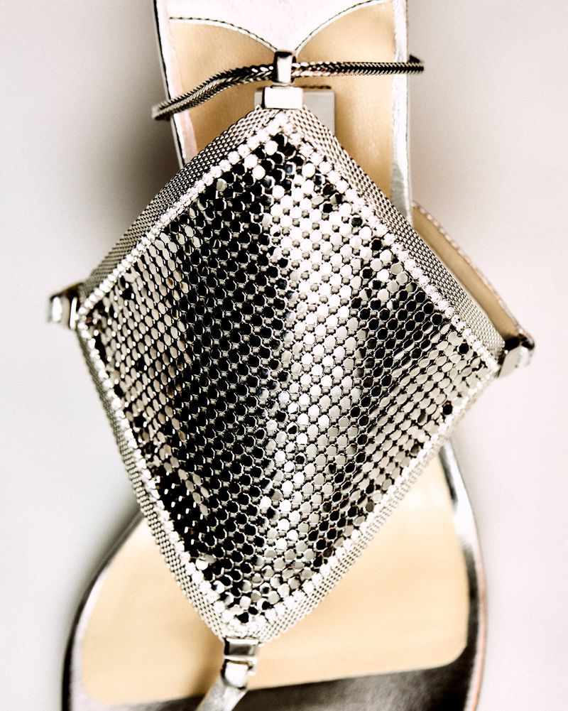 Close-up of a silver sandal with a diamond-shaped metallic mesh decoration, set against a neutral background.