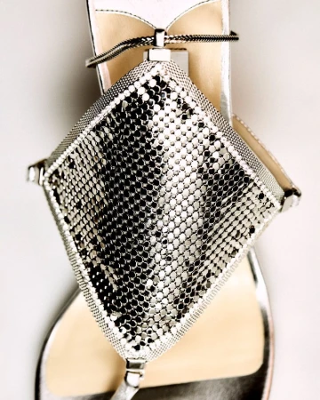 Close-up of a silver sandal with a diamond-shaped metallic mesh decoration, set against a neutral background.