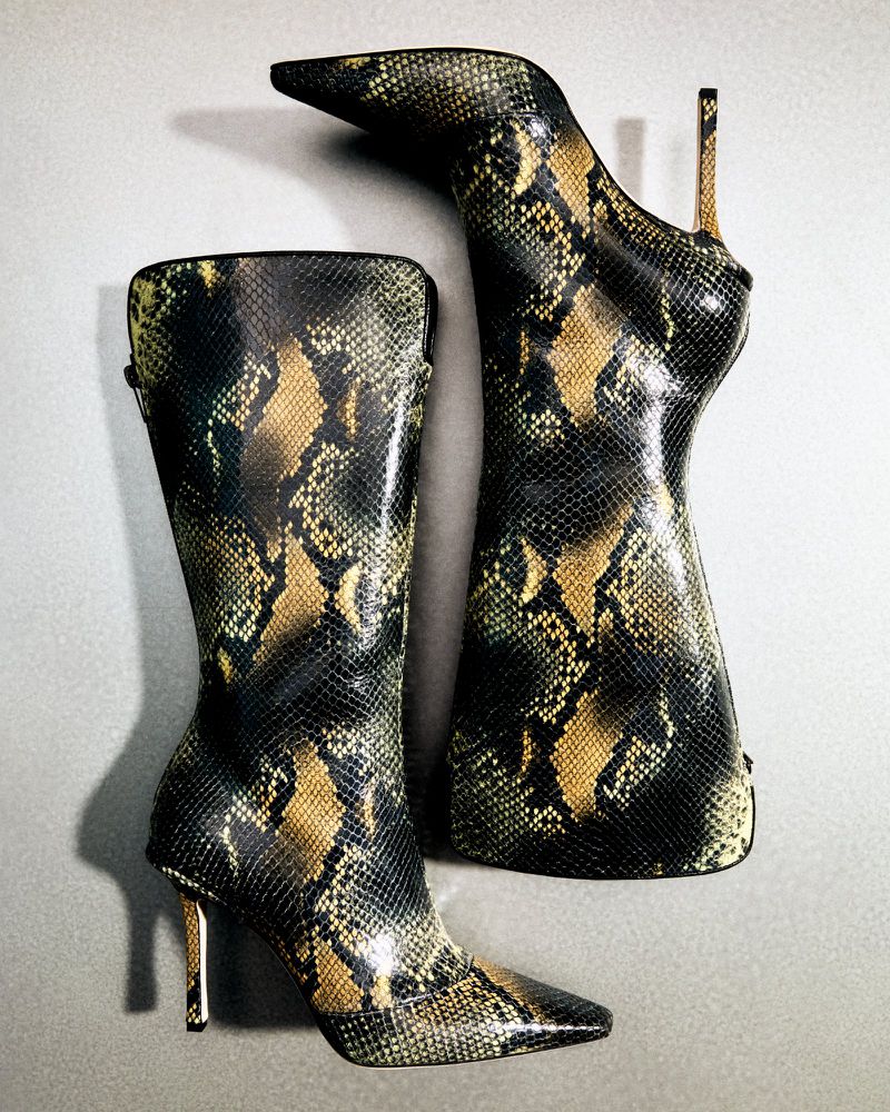Two snake-print knee-high heeled boots are displayed on a neutral surface, one upright and one angled.