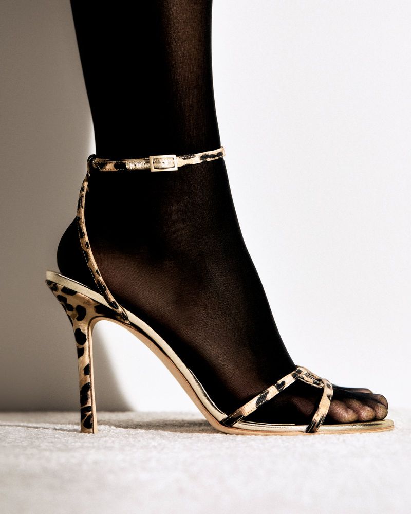 Close-up studio shot of a woman's foot wearing black tights and a high-heeled sandal with an animal print strap.