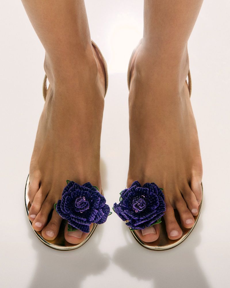 Woman's bare feet in stylish gold sandals adorned with purple flower embellishments against a seamless white backdrop. Fashionable footwear.