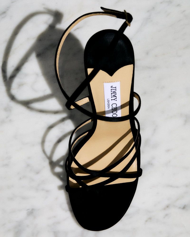 Elegant black high-heel sandal with a multi-strap design against a white marble backdrop, showcasing its sleek style and luxury brand label.