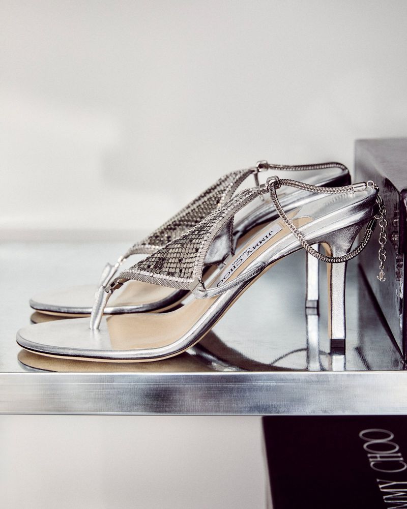 Jimmy Choo silver strappy heeled sandal on a shelf, close-up fashion product shot with bright lighting and a clean, modern setting.