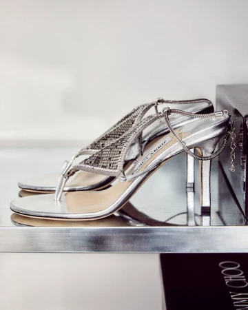 Jimmy Choo silver strappy heeled sandal on a shelf, close-up fashion product shot with bright lighting and a clean, modern setting.