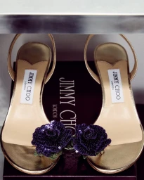A pair of gold slingback sandals featuring dark purple sequined rose embellishments resting on a branded Jimmy Choo shoebox.