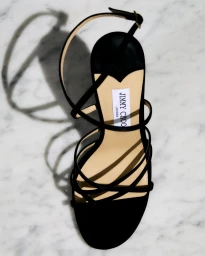 Top-down view of a black strappy Jimmy Choo high heel shoe on white marble surface casting a distinct shadow.