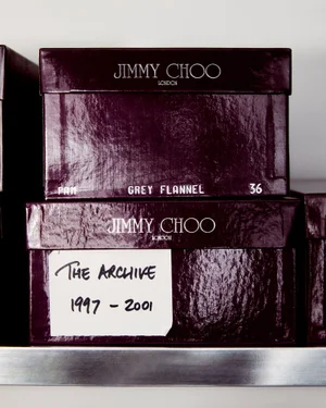 Two dark purple glossy Jimmy Choo shoeboxes stacked on a metal shelf, with handwritten archival labels.