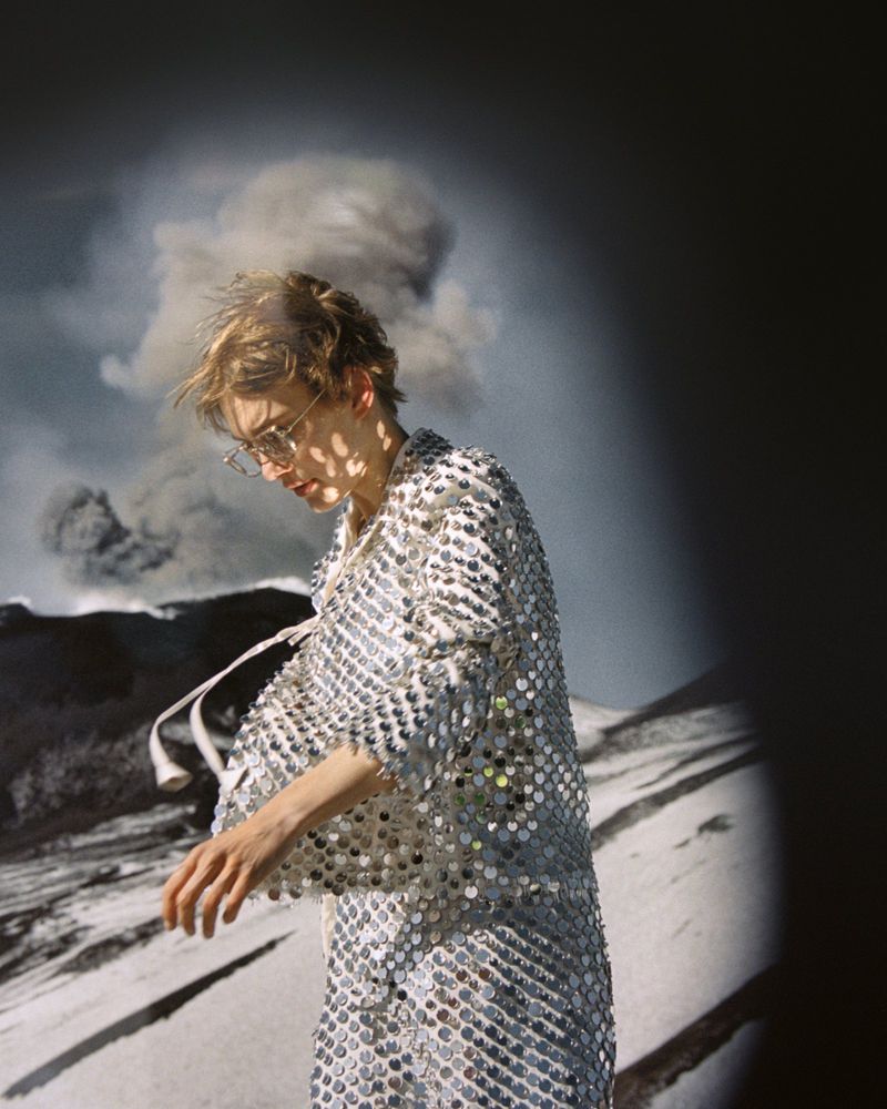 Adult woman wearing shiny silver coat poses in front of volcanic landscape with smoke and ash cloud in an outdoor fashion shot.