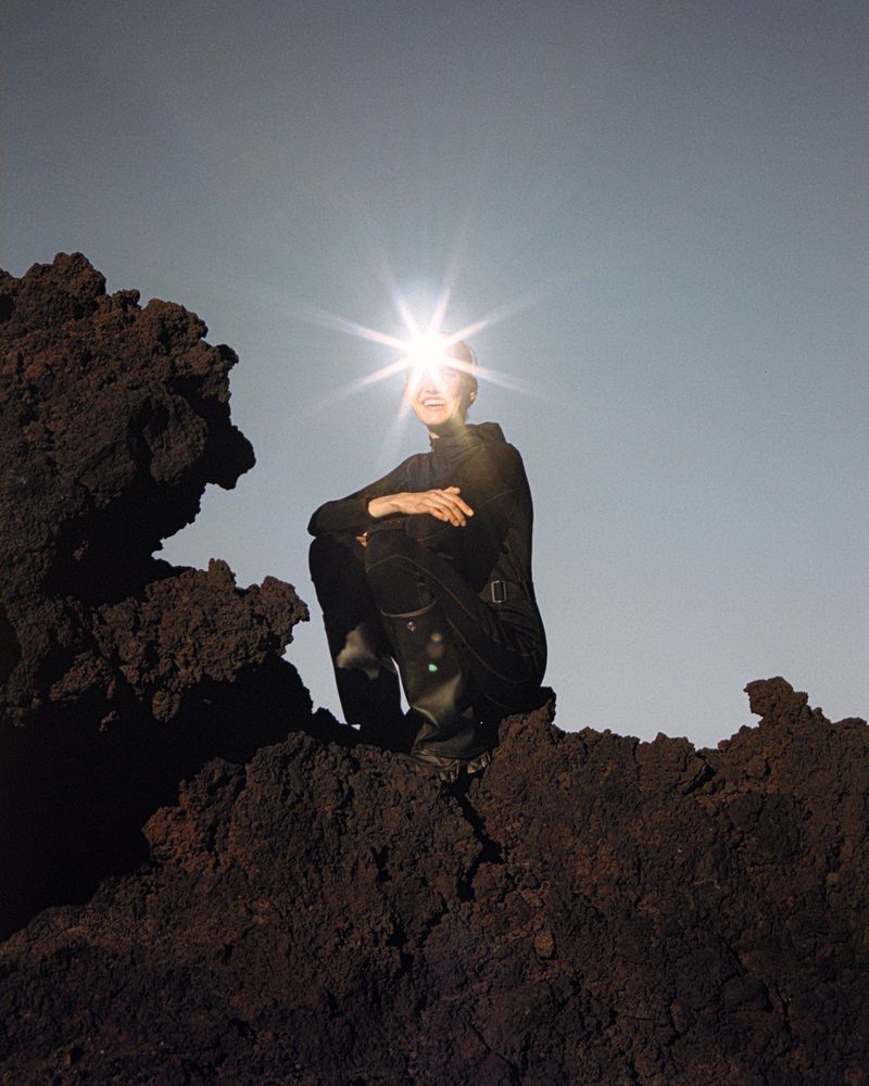 Adult woman crouches on dark volcanic rock under direct sunlight against pale sky. She wears dark clothing and smiles brightly.