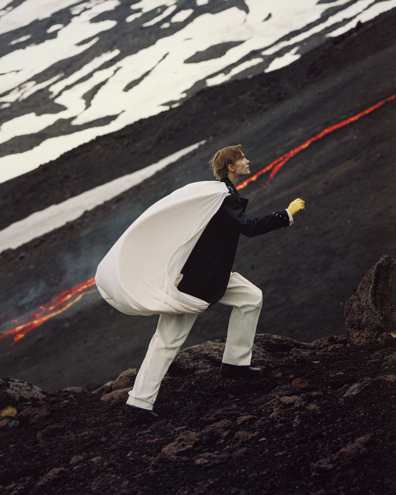 A model in white pants and dark jacket walks up a rocky slope with streaks of lava and snow, carrying a large white fabric bag.