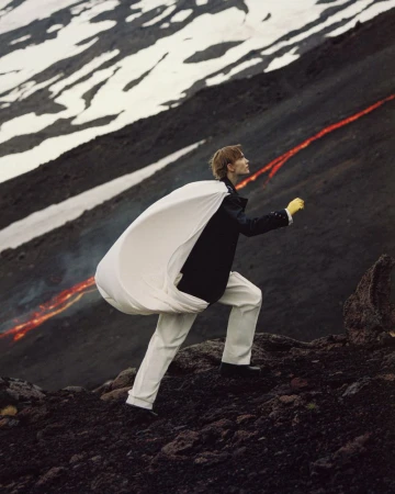 A model in white pants and dark jacket walks up a rocky slope with streaks of lava and snow, carrying a large white fabric bag.