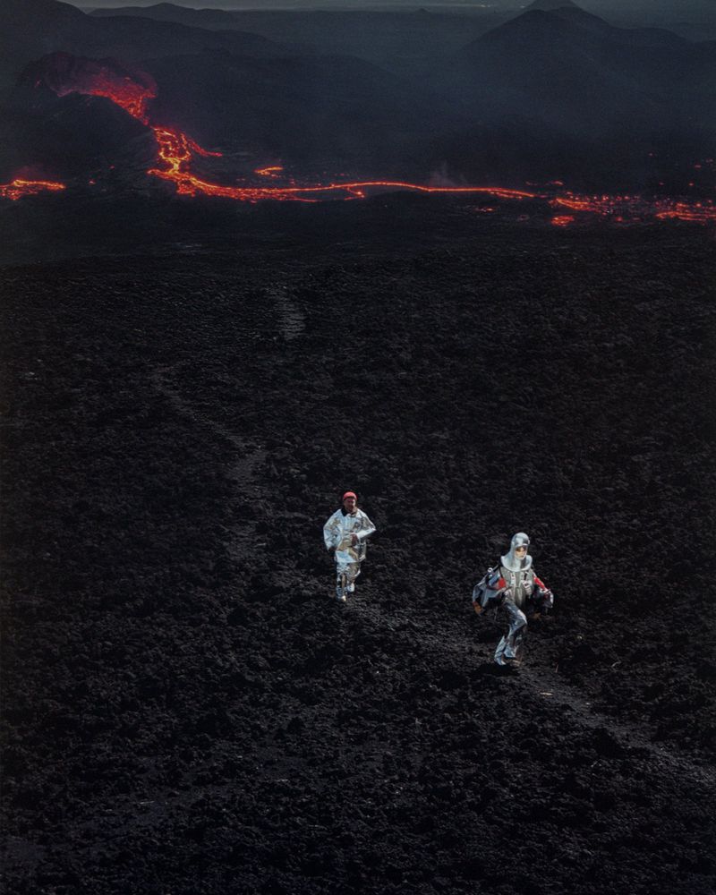 Two people in protective suits run across volcanic landscape. Molten lava river in background. Dark, dramatic photography.