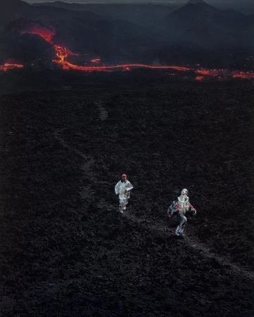 Two people in protective suits run across volcanic landscape. Molten lava river in background. Dark, dramatic photography. 