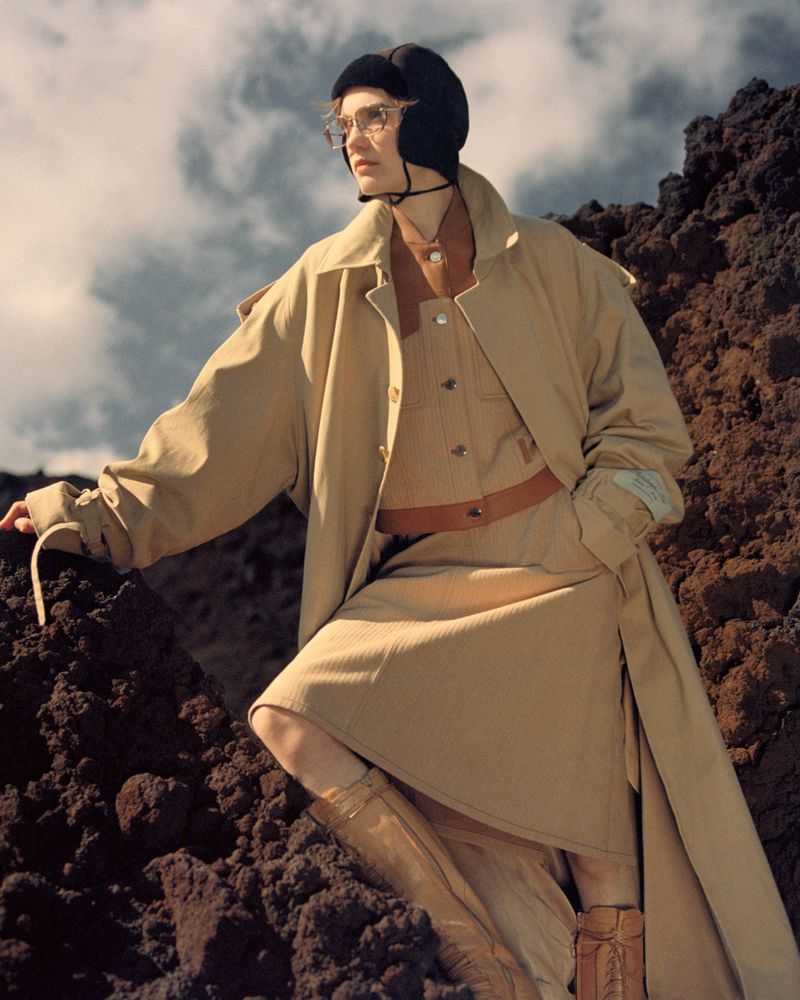 Woman in a tan trench coat, glasses, and a black aviator hat posing on a rocky landscape with a cloudy sky in an outdoor fashion shoot.