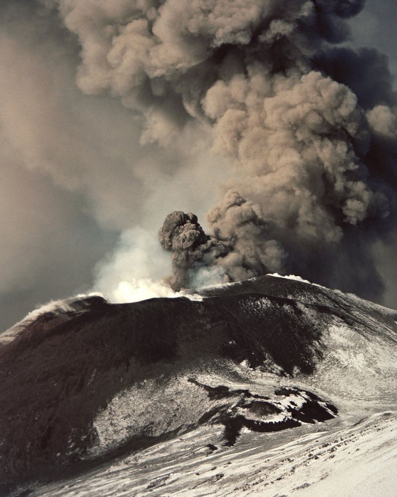 Volcano erupting, spewing thick smoke and ash into a cloudy sky, with snow-covered slopes visible in the foreground of a natural disaster scene.