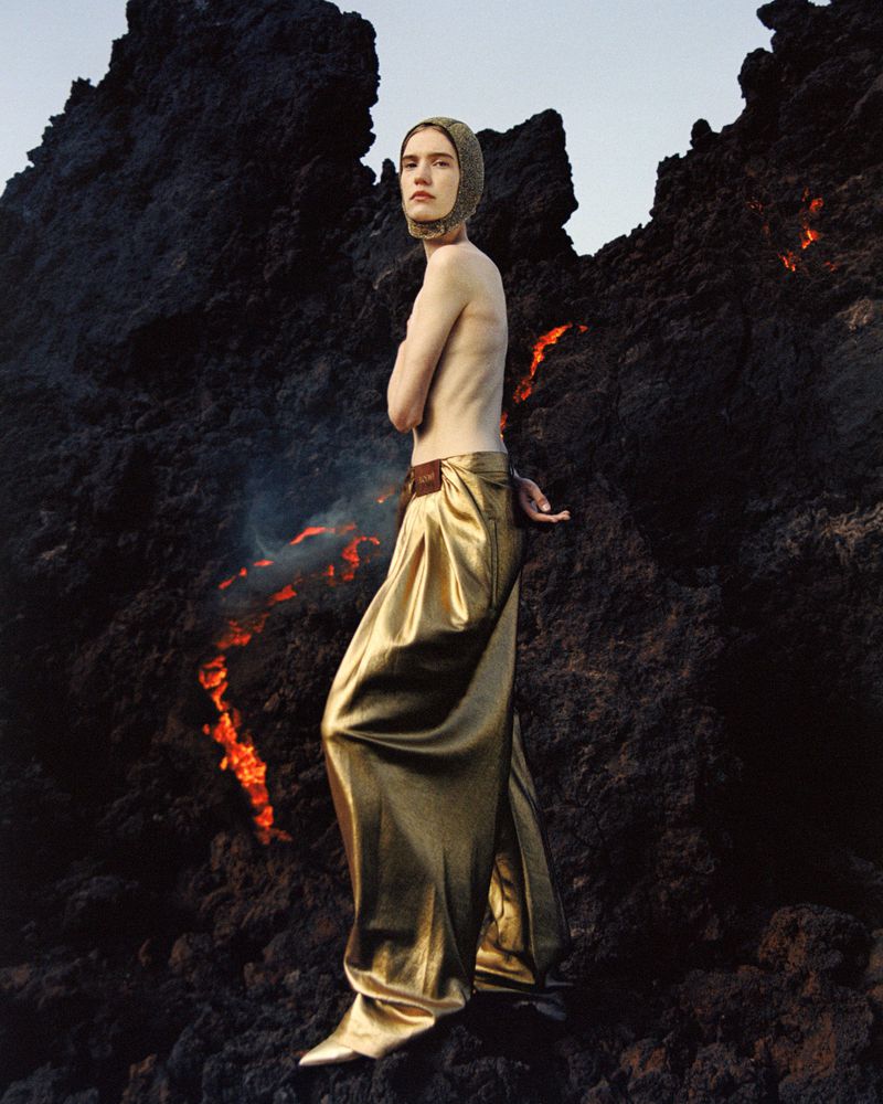 Adult fashion model poses in gold trousers on a black lava field with fire. The sky is light and the light is bright and soft.