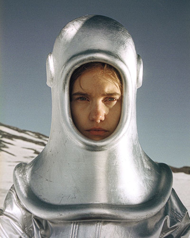 Portrait of a fair-skinned woman in a silver space helmet and matching suit against a snowy landscape under a pale blue sky.