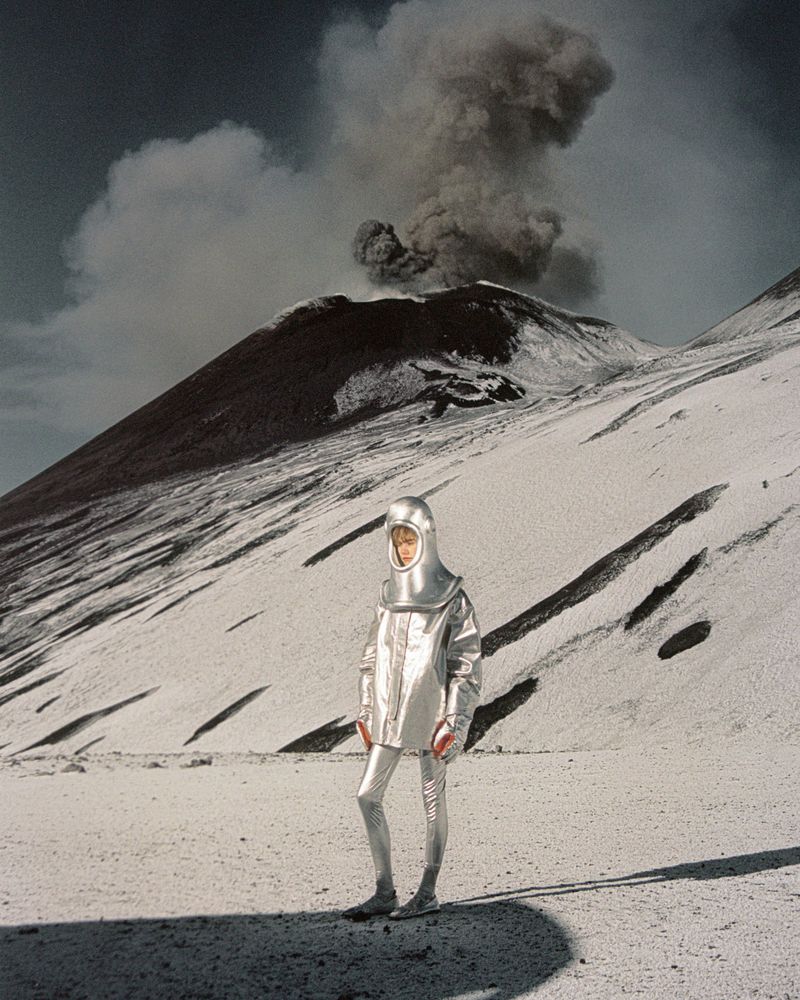 Person in a silvery protective suit stands on a white surface, against the backdrop of a volcano emitting smoke under a daytime sky.