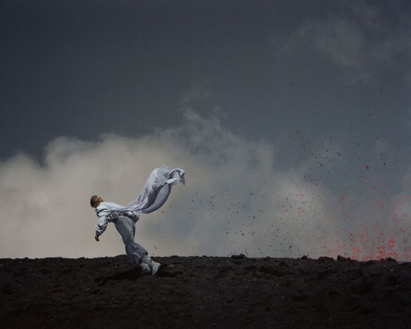 Person in a white outfit and fabric falling backwards on a volcanic landscape with lava, dark clouds, and a dramatic sky.