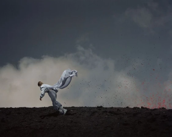 Person in a white outfit and fabric falling backwards on a volcanic landscape with lava, dark clouds, and a dramatic sky.