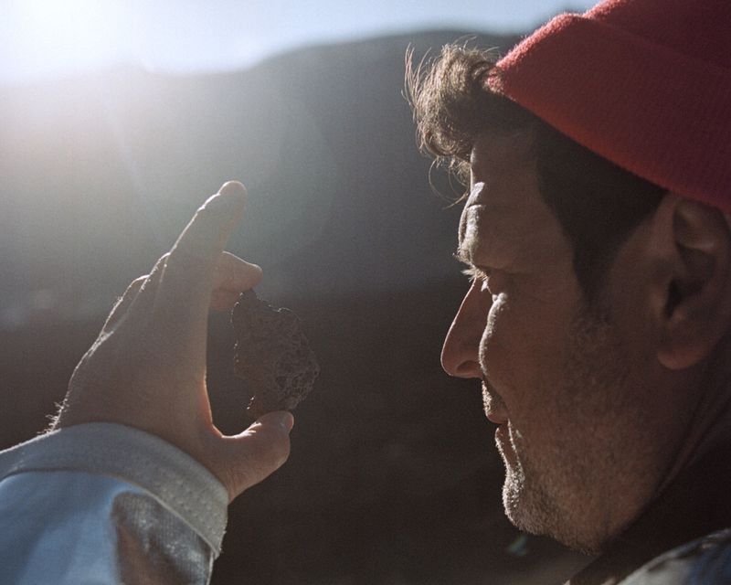 Adult male in red beanie holds dark mineral rock outdoors in bright sun. Close-up profile highlights mineral and man in natural setting.