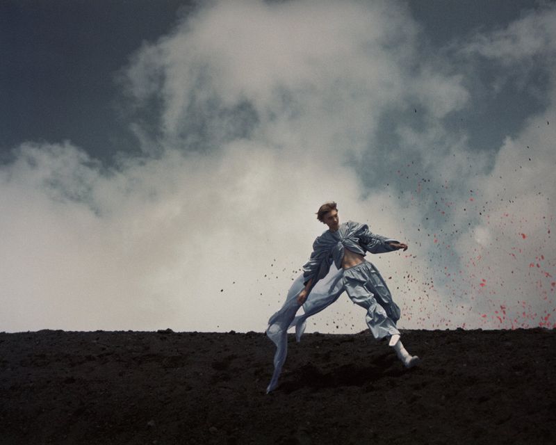 Fashion shot of androgynous adult in a light blue outfit posing on a black landscape with a cloudy sky in the background.