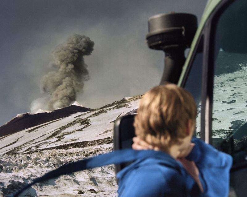 A person looks out the window of an SUV at a snow-covered volcano erupting under a cloudy sky in a landscape vista.