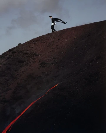 Adult male in dark suit walks down volcanic slope with lava stream. Dramatic landscape and unusual fashion-forward editorial concept.